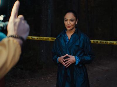 Kvinde i mørk frakke poserer foran et kamera med en politiafspærring bag hende.